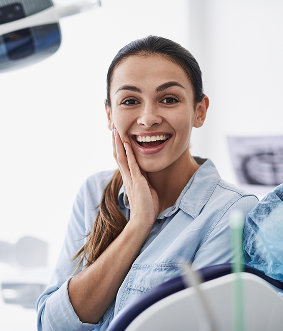 A woman is smiling in a dental office, with her hands on her cheeks, seated in front of a dental chair.